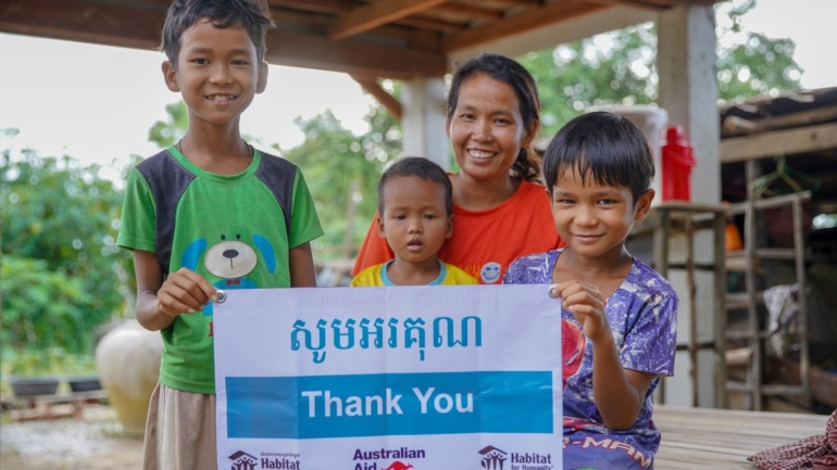 A Cambodian woman and her three children holding a Thank You sign in front of their new home.