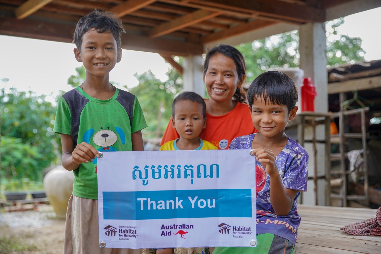 A Cambodian woman and her three children holding a Thank You sign in front of their new home.