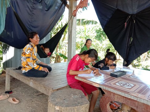 A woman and older woman sit on a raised platform as they watch two girls doing schoolwork at a wooden table. A young boy is lying in a hammock just behind.