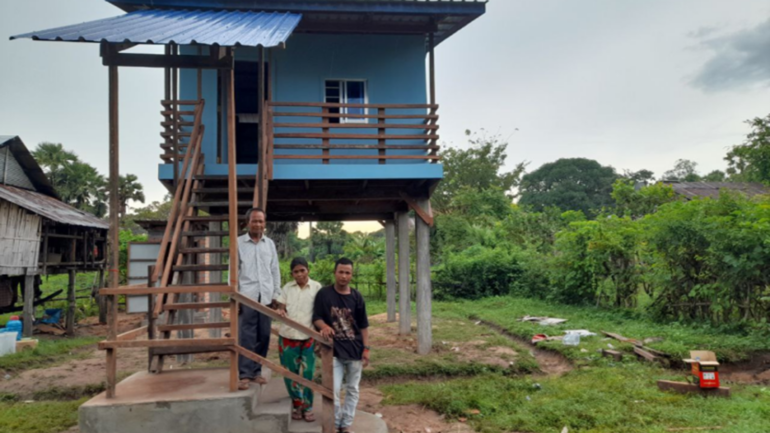 A father, mother and son standing on the stairs leading up to their blue house on stilts.