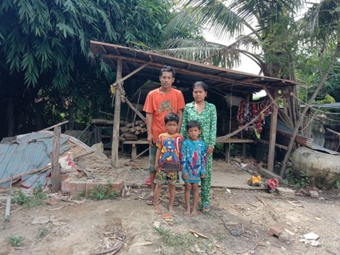 A family of four stand in front of a delapidated hut where they used to live.