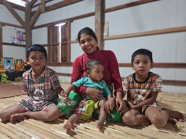 A woman sits with her three sons inside their new home. She's smiling as she holds her youngest child.