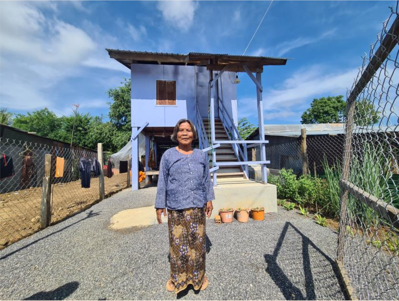Yip Say, an elderly Cambodian woman, in traditional dress, stands proudly in her gravel driveway, outside her new home. The home is painted blue which matches the sky behind it.