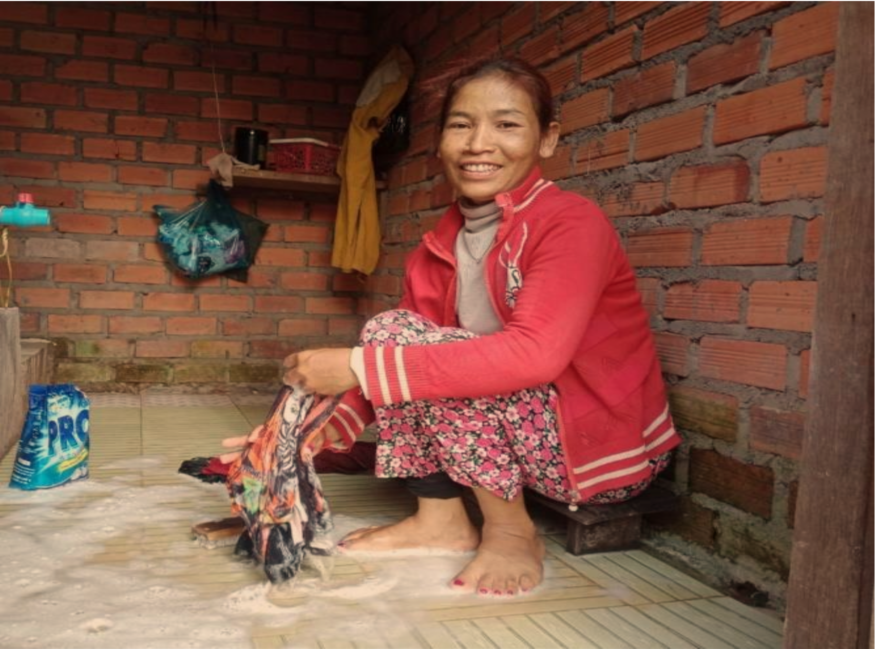 A smiling woman, Sophay crouches on the soapy, tiled floor of her new washroom, doing her laundry.