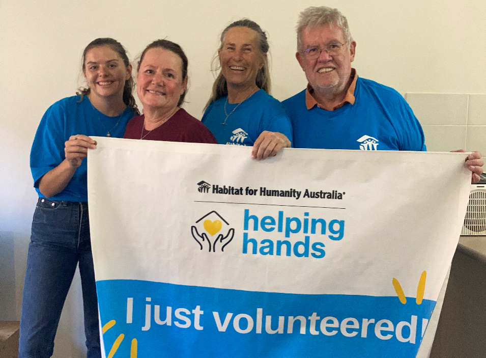 Three women and one man, stand in a line, against a blank white wall. They are holding up a large banner that reads 'Habitat for Humanity Australia. Helping Hands: I just volunteered!' 