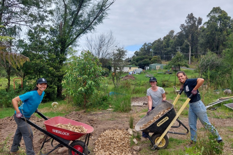 three women stand to the left and right of a pile of mulch. They all hold wheelbarrows and are smiling toward the camera.