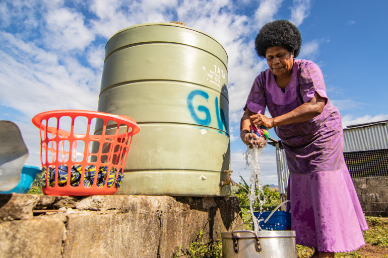 A Fijian woman wrings out her washing over a silver pot. She stands, outside, next to a water tank and an orange basket.