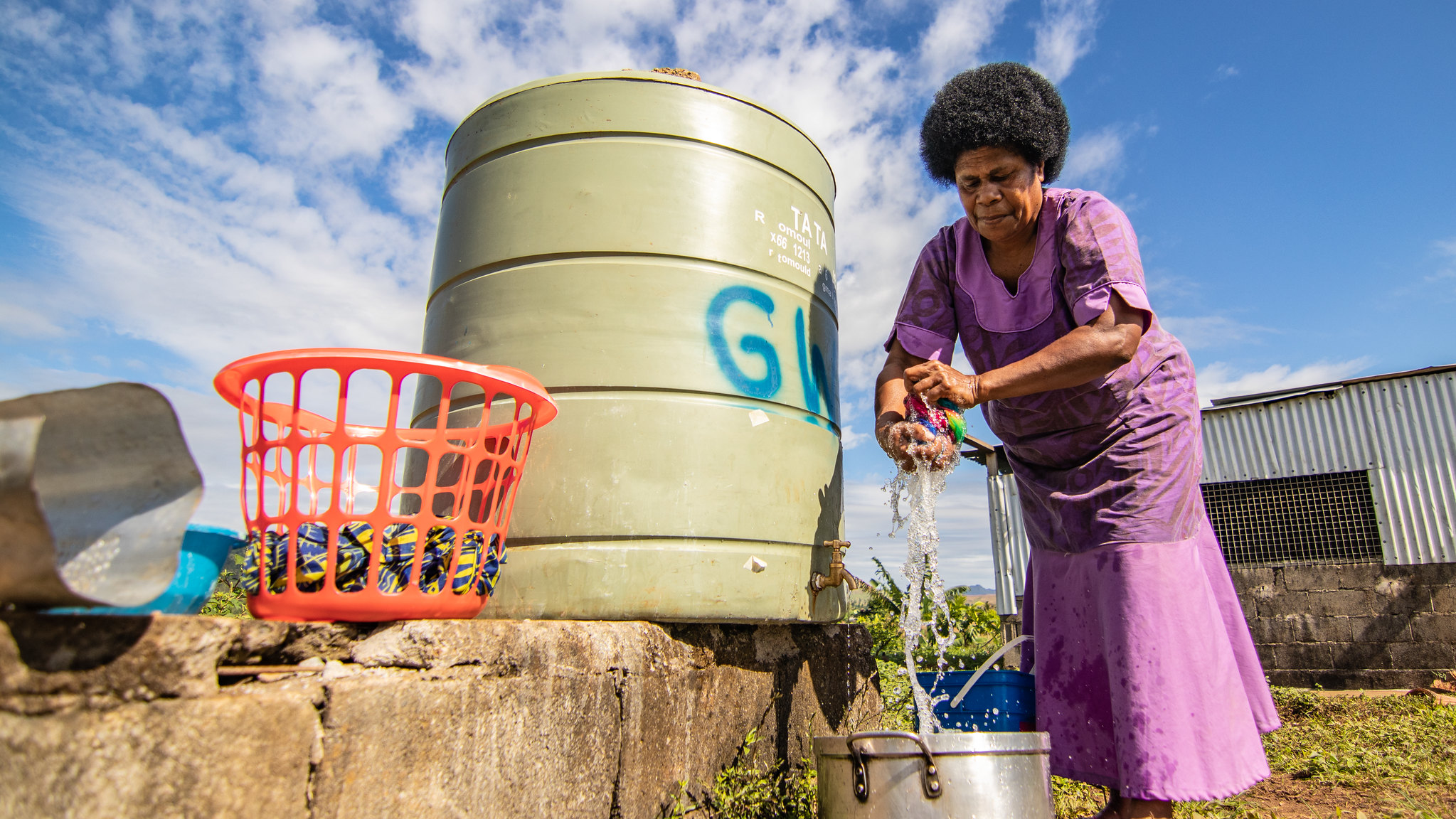 A Fijian woman wrings out her washing over a silver pot. She stands, outside, next to a water tank and an orange basket.