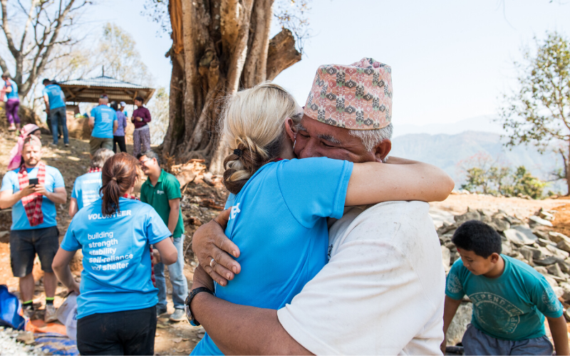 A female volunteer in a blue Habitat for Humanity t-shirt hugs a Nepalese man wearing a traditional hat. The image shows the joy of coming together to support each other. 
