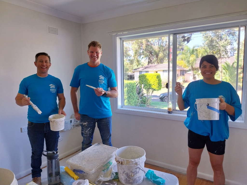 Three Habitat Volunteers, wearing blue Habitat t-shirts stand in the corner of a room, holding buckets of white paint and paint brushes as they volunteer on the Brush with Kindness program

