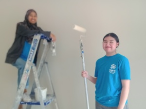 Lavinia stands in the foreground holding a paint roller, a fellow volunteer stands on a ladder behind her