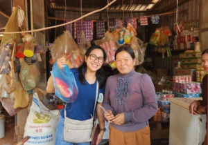 Two women stand at a local shop. One woman lifts a plastic shopping bag up. The shop sells the bottled water from the water station.