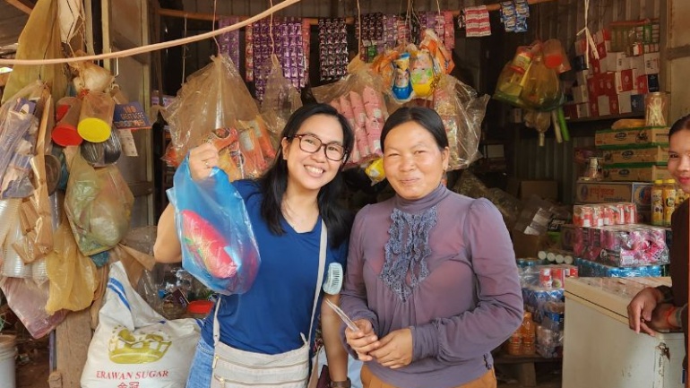 Two women stand at a local shop. One woman lifts a plastic shopping bag up. The shop sells the bottled water from the water station.