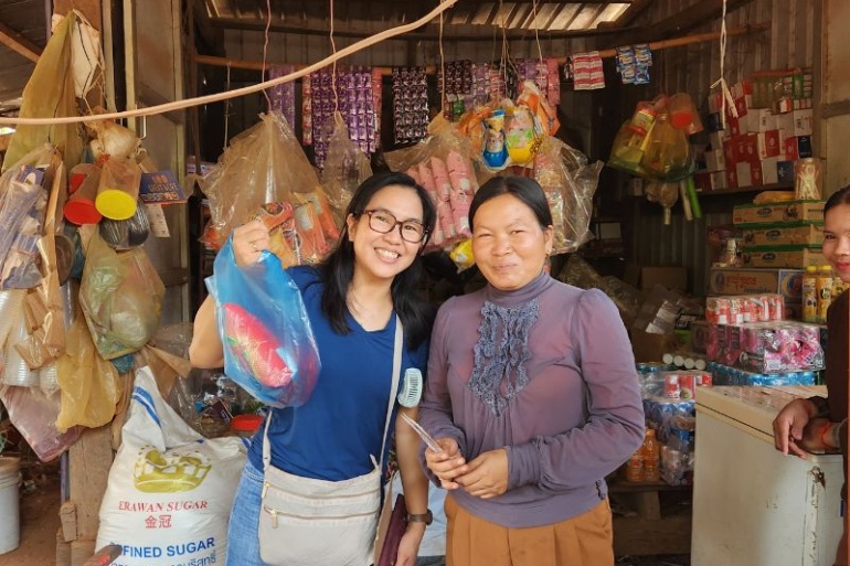 Two women stand at a local shop. One woman lifts a plastic shopping bag up. The shop sells the bottled water from the water station.