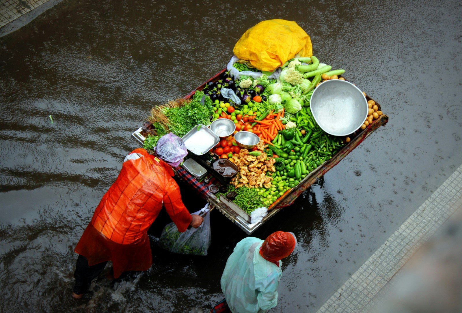 two men push a wooden cart of fresh produce through flood water